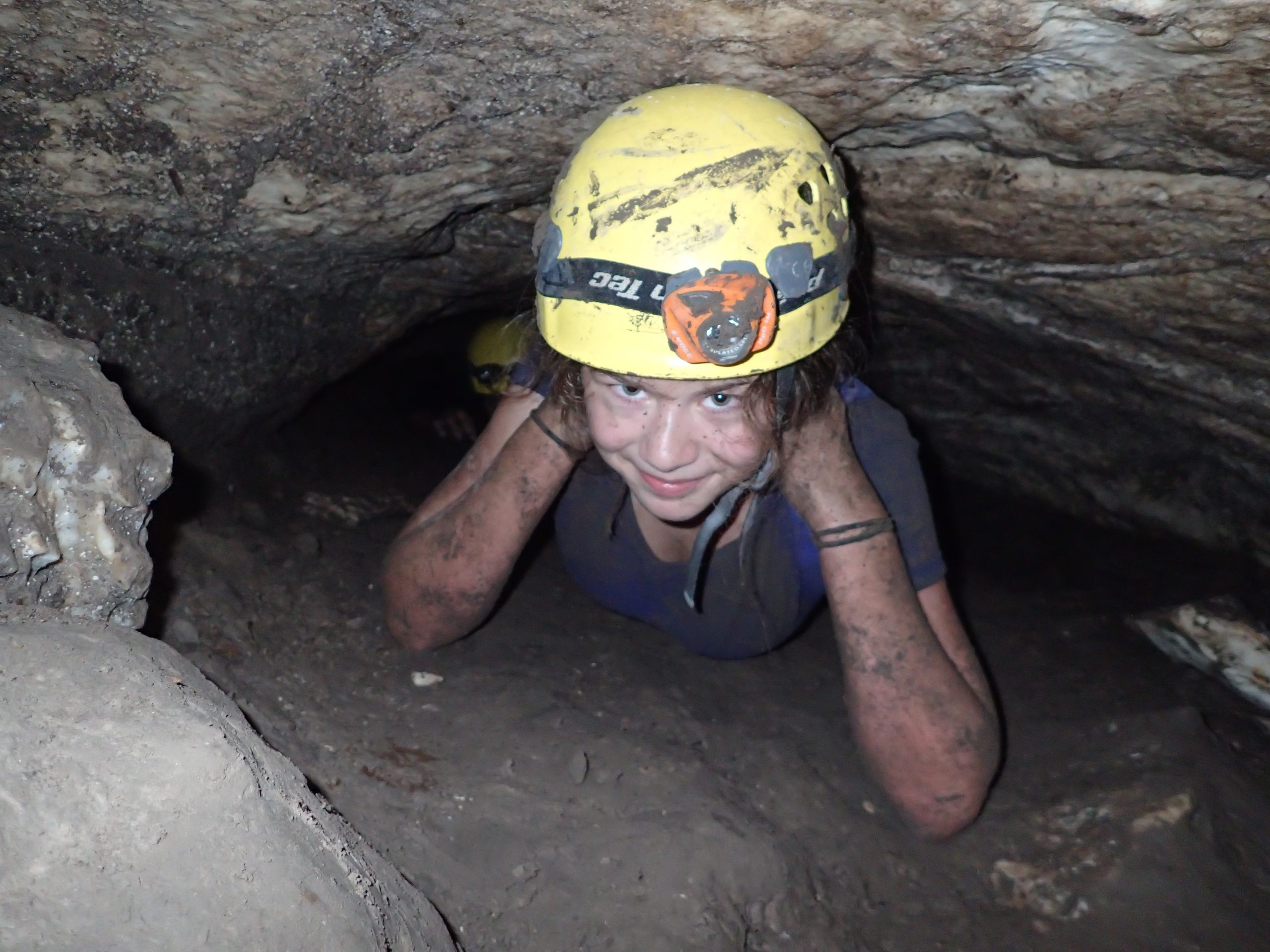 Kid in a cave covered in mud wearing a yellow helmet with a headlamp