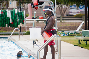 A lifeguard watching over a pool. He is wearing a white visor, has a whistle in his mouth and is holding a red lifeguard float.