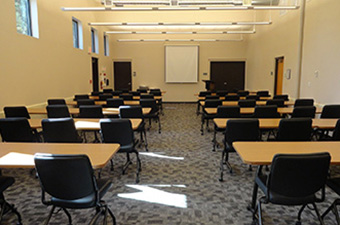 A large community room with 13 long tables and chairs, with a projector screen on the wall