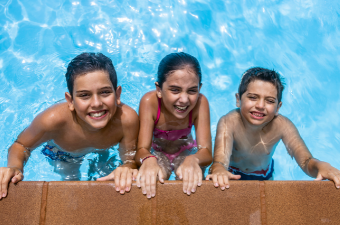 three kids on smiling while holding the side of the swimming pool